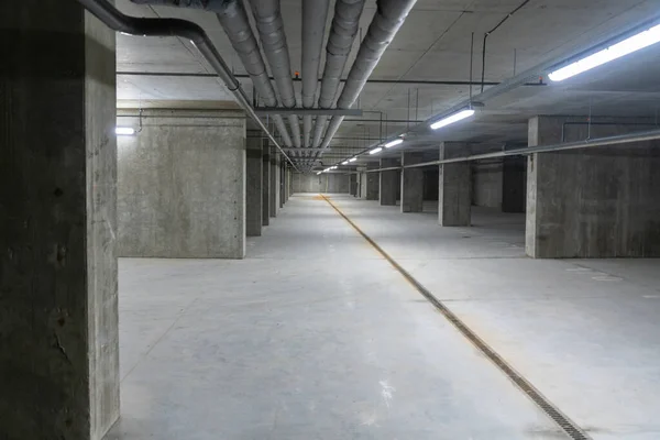 セメント廊下に沿って後退するビューで灰色のコンクリート商業地下に露出した天井のパイプや側面へのサポート柱とネオンライトで照らされたドライブウェイ — ストック写真