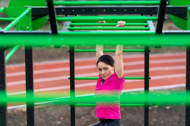 Açık hava spor sahasında paralel barlarda çalışan genç bir kadın sağlık ve fitness konseptinde ekipmanların arasından izleniyor.