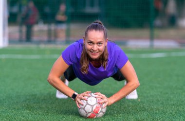 Genç bir kadın, kameraya gülümserken kafasında bir topla spor sahasında plantasyon egzersizleri yapıyor.