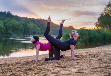 İki zarif genç kadın senkronize yoga yapıyor. Gün batımında kumlu bir sahilde bacaklarıyla poz veriyorlar. Sağlık ve zindelik ya da aktif yaşam tarzı konseptiyle.