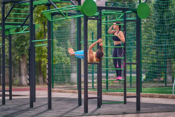 Young man and woman working out outdoors on open air gym equipment using the parallel bars in a leafy green park or sporting facility in a health and fitness concept