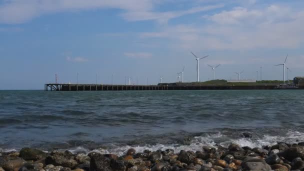 遠くに風力タービン ビーチ石 桟橋や産業海岸破砕海波のローアングル ショット — ストック動画