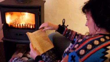 Sıcak kazak giyen kadın ateşin yanında oturan ve bir kitap okuma. Şömine üzerinde odaklanmak