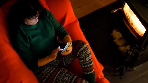 ニットのセーターと暖炉と彼女の携帯電話やスマート フォンで入力テキスト メッセージによって居心地の良い赤いソファーに座っていたレギンスを着て直接上記の女性観 — ストック動画