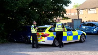 SALFORD, MANCHESTER, İngiltere - Mayıs 2020: İkamet alanındaki küçük bir otoparkta şehir caddesi. İki İngiliz polis memuru ağacın altında şüpheli bir BMW 'nin yanına park etmiş polis arabasının yanında konuşuyor..