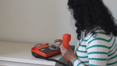 Beyaz masa ve duvarın yanında oturan ve eski turuncu kahverengi renkli sabit hat üzerinde çeviren uzun siyah kıvırcık saçlı kadının yan görüntüsü..