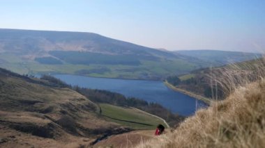 İngiltere, İngiltere 'de Peak District Ulusal Parkı' nda güneşli bir günde Dove Stone Reservoir 'un üzerindeki güzel bir manzarada kırmızı ceket giyen şehir kadını tepeye tırmanıyor..