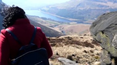 İngiltere 'nin Peak District Ulusal Parkı' ndaki Dovestone barajının yukarısındaki kayanın etrafındaki tepeden aşağı doğru yürüyen kırmızı ceketli kadının el kameralı görüntüsü..