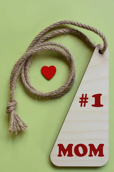 #1 mom - text on wooden medal with red little heart hanging on natural linen twine on light green background top view. Family and love concept.