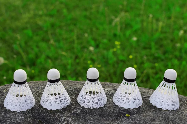 Five white plastic badminton shuttle cock on grey stone on green grass background with copy space for text on blurred grass.