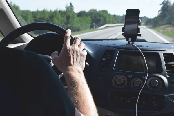 Man hand holding car steering wheel inside cabin with multimedia system, black dashboard and smartphone for navigation with road and green trees view from window.