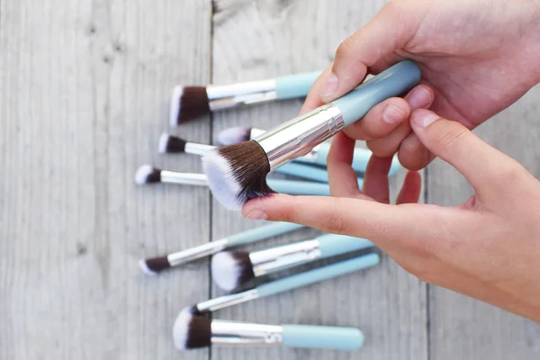 Hands holding makeup brushes set on grey weathered wooden table background with copy space for text. Beauty visage and makeup design concept.