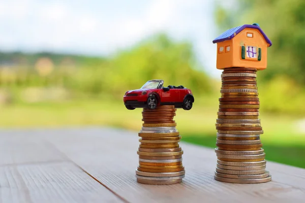 House and car on money coins stack on wooden table outdoors on green blurred background with copy space for text. Saving to buy house or new car. Property investment and money saving financial concept.