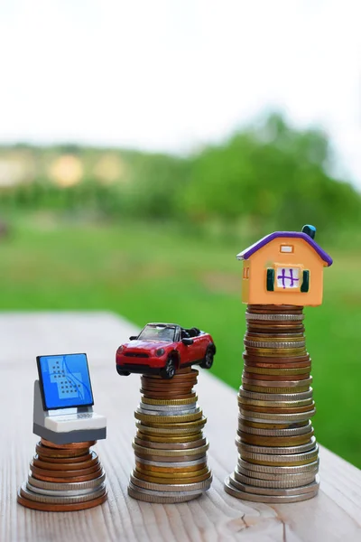 Computer car house on coins stack on wooden table on green blurred background outdoors with copy space. Saving to choose and buy necessary goods or property. Different appropriate amount  investment and money saving financial concept.