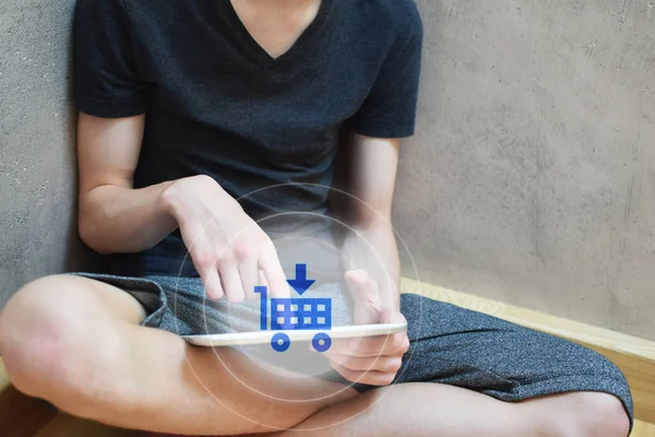 Young man sitting on floor using tablet and pressing transparent blue white virtual interface button with cart icon on grunge light brown background with copy space for text. Online shopping, add to cart, purchasing and selling business concept.