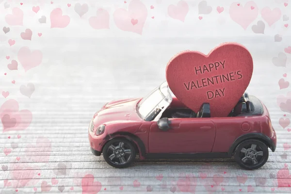 Red toy car delivering Valentine's day heart on blurred wooden background with copy space for text or message greeting card illustrated transparent hearts around.