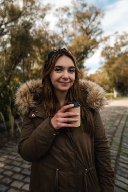 Parkta elinde kahve tutarken kameraya gülümseyen beyaz bir kadın.