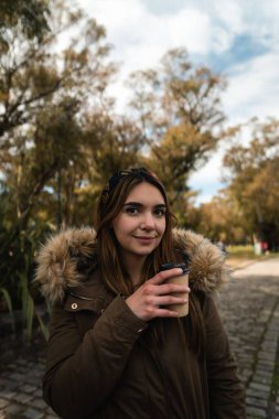 Kahve içerken gülümseyen ve parkta yürüyüş yapan beyaz bir kadın. Dikey