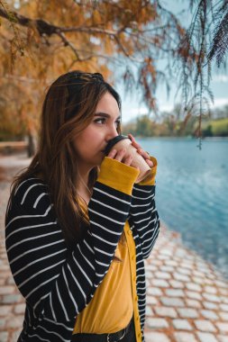 Güz parkında göle bakarken kahve içen beyaz bir kadın. Dikey