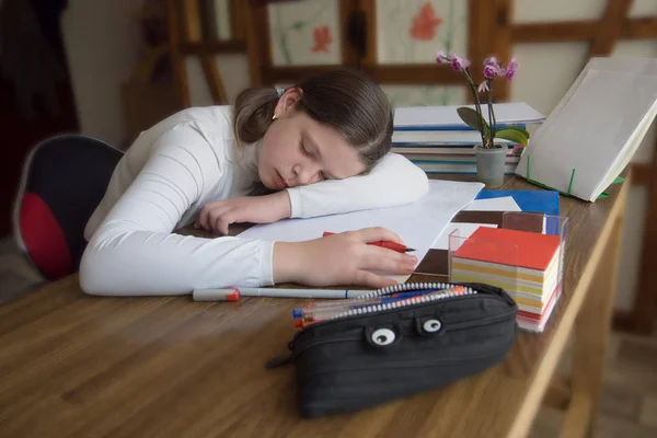 Teenager girl sleeping tired at the table during homework with a pen in hand, on the table there are a pencil case with pens, stand for books, notebooks, a stack of schoolbooks and an orchid in a pot
