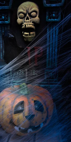 Halloween pumpkin Jack of the Lantern with illuminated eye sockets,black raven and tombstone rest in peace with skull on it,covered in cobwebs.Vertical banner,selective focus.Scary Halloween concept — Stock Photo, Image