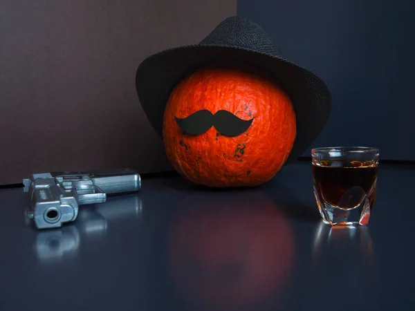 Halloween pumpkin as a man with moustache,wearing black hat on dark background,grey pistol and shot glass of spirits.Concept of trick or treat,aggression,armed protest,complex relationships.Copy space — Stock Photo, Image