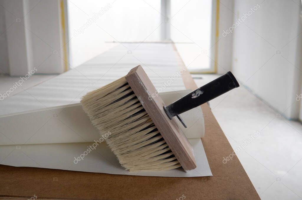 Wide paint brush for glue with wooden base for bristle,black handle,several sheets of white wallpaper unfolded in length on table for pasting against white wall,floor-to-ceiling French window in room.