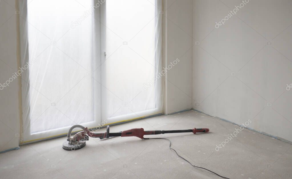 Dirty and dusty red sander for walls and ceilings lies on the floor in empty room against white ready-to-paint walls and floor-to-ceiling French window.Concept of repair of apartment,building house.