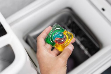 Çamaşır deterjanı kapsülünü çamaşır makinesine koyan adam, yakın plan. Kapsülde renkli çamaşır ekosu jeli. Çamaşır yıkıyorum. Yıkama ve temizleme kavramı
