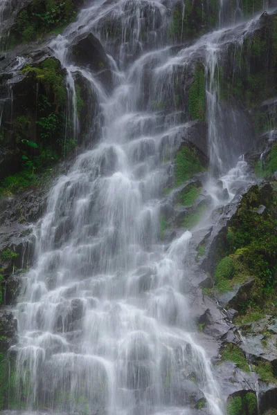 A natural waterfall flows down a long line from a cliff into a sprinkling valley.
