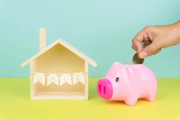 saving money to buy a house concept, hand putting a golden coin into piggy bank and wooden house model with sale sign