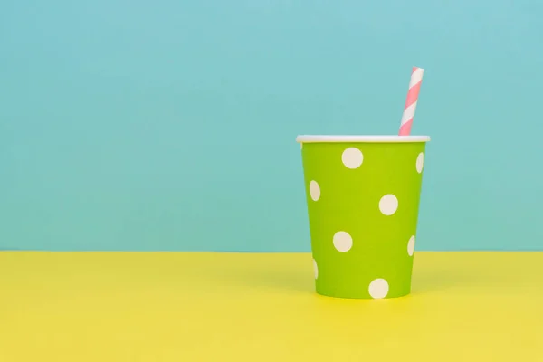 a green polka dot party paper cup with pink striped straw on the yellow table with blue background
