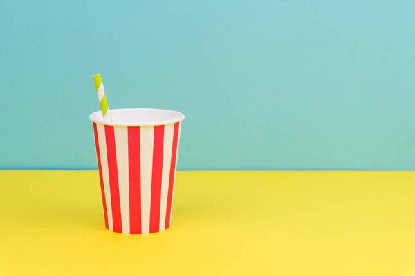 полосатая бумажная чашка с зеленой полосатой соломой на желтом столе с синим фоном
