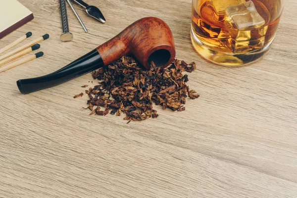 a smoking pipe with tobacco, pipe tamper tool, matches and a glass of whisky on wooden table