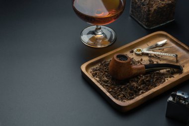 bir sigara boru ve boru dış müdahale aracı içinde ahşap tepsi, bir krom daha hafif, bir bardak brendi siyah masada tütün kavanozu