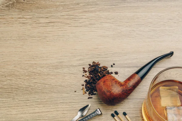 a smoking pipe with tobacco, pipe tamper tool, matches and a glass of whisky on wooden table