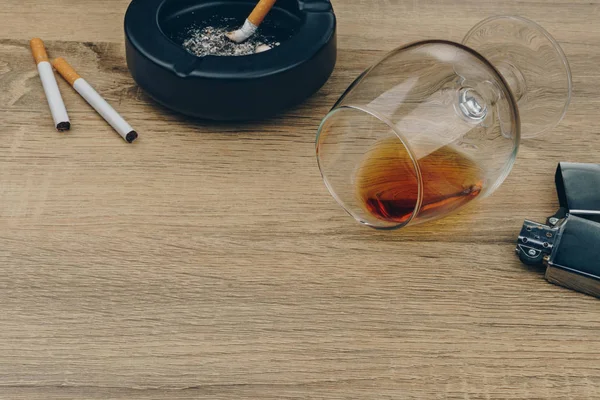 a glass of brandy, cigarettes, metallic lighter and black ceramic ashtray full of ashes with the cigarette butts on the wooden table