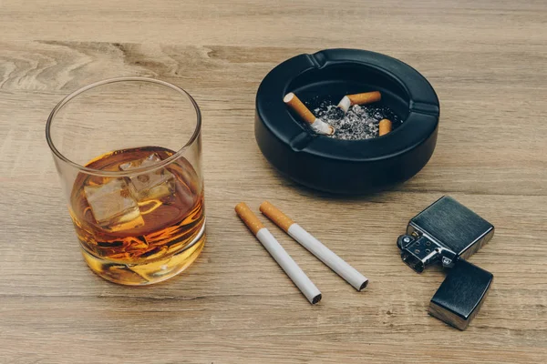 a glass of bourbon whiskey, cigarettes, metallic lighter and black ceramic ashtray full of ashes with the cigarette butts on the wooden table