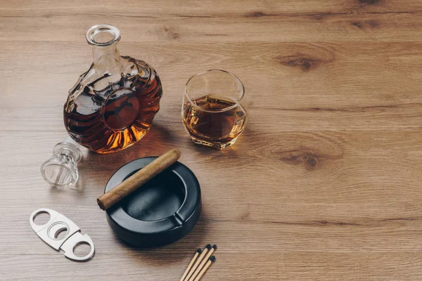 Cuban cigar on a black ceramic ashtray, a glass of bourbon whiskey with a whiskey decanter, a stainless steel cigar cutter and matches on the wooden table