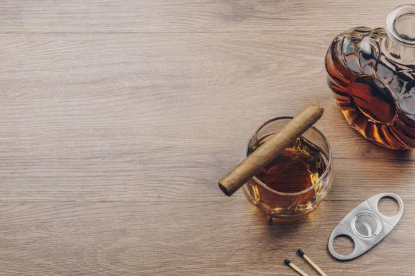 Cuban cigar on a glass of bourbon whiskey, a whiskey decanter with a stainless steel cigar cutter and matches on the wooden table