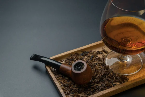 close up of a smoking pipe with tobacco and a glass of brandy in wooden tray