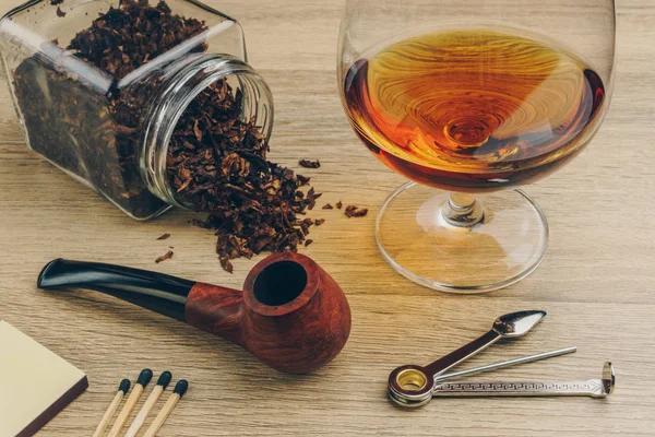 a smoking pipe with tobacco jar, pipe tamper tool, matches and a glass of brandy on wooden table