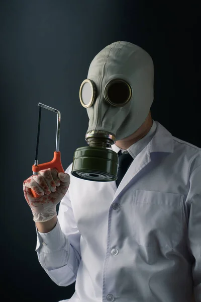 Evil and creepy medical experiment concept, a scary doctor in gas mask wearing bloody gloves with a hand saw on dark background