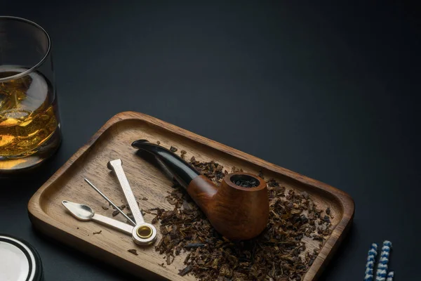 a smoking pipe with tobacco and a tamper tool in wooden tray, a glass of bourbon whiskey and pipe cleaners on the black table