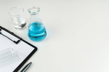laboratuvar aletleri. Mavi bir sıvı şişesi, berrak sıvı kabı, test tüpleri ve beyaz masaya kalemiyle bir pano