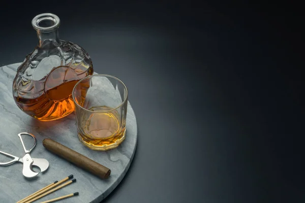 a Cuban cigar and a stainless steel cigar cutter, a glass of bourbon whiskey with a whiskey decanter and matches on the marble tray