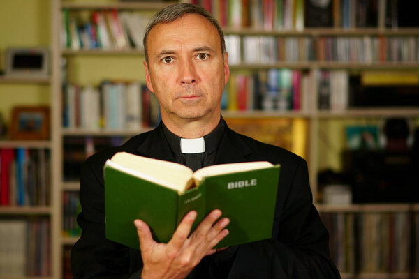 Хорошо выглядящий серьезный католический священник изучает Библию в своей библиотеке. Он смотрит на нас.
.                               