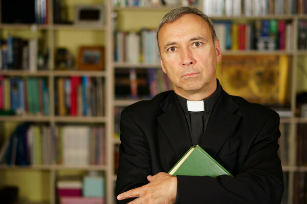 Красивый католический священник учится в своей библиотеке. Он смотрит на нас с интересом, неодобрением и осуждением
.                               