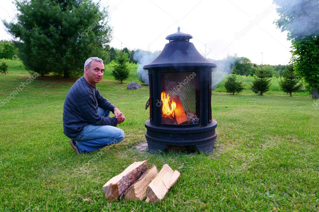 A caucasian man, 50's, just lit his outdoor fireplace, during evening, in his rural backyard.
