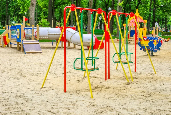 緑の木々に囲まれた公園でカラフルな子供の遊び場活動。子供たちは、現代の遊び場で走り、滑り、スイングします。都市部の子供時代の概念. — ストック写真